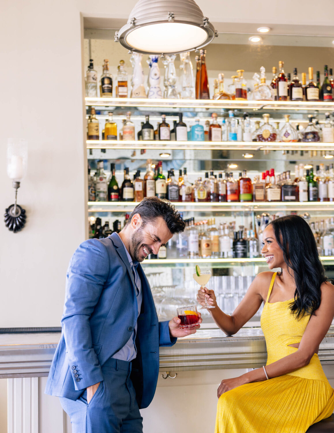 A man and woman in formal attire enjoying drinks and laughing at a bar with liquor shelves.