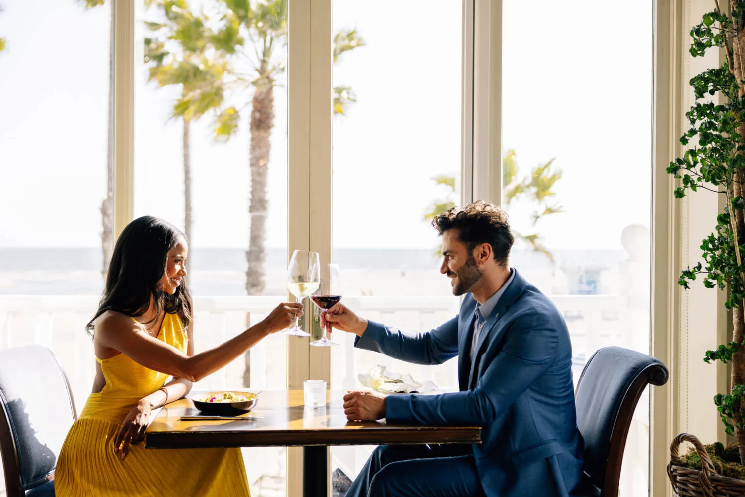 A couple toasting wine glasses at a table by a window with a view of palm trees.