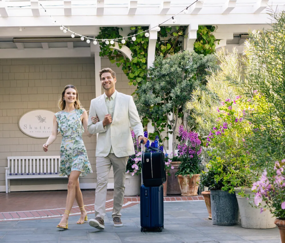 Smiling couple arriving at a hotel entrance with greenery, flowers, string lights, and luggage.