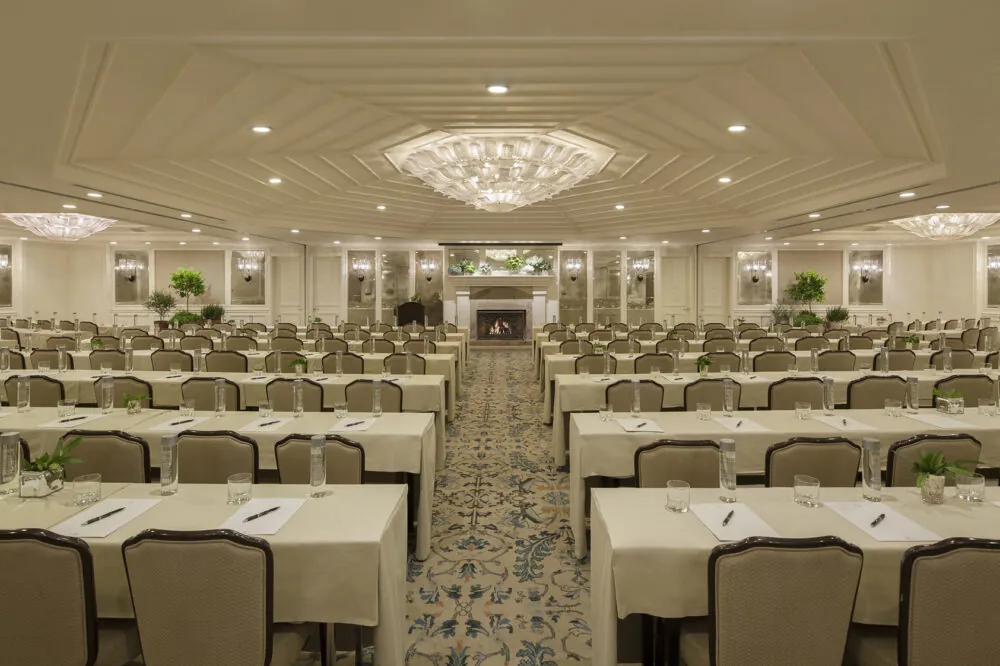 A grand conference room with elegant chandeliers, neatly arranged tables, and a warm fireplace.
