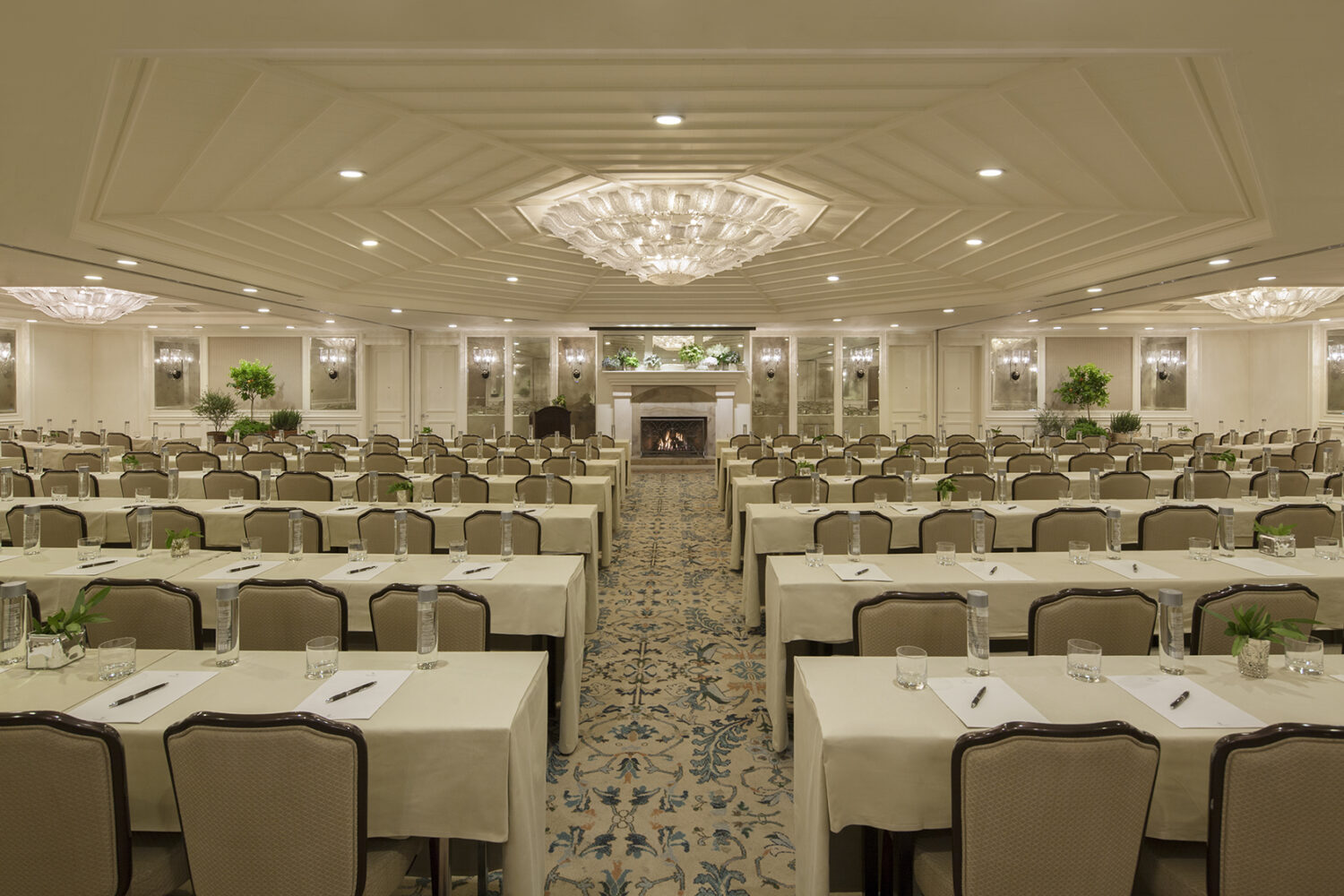 A grand conference room with elegant chandeliers, neatly arranged tables, and a warm fireplace.