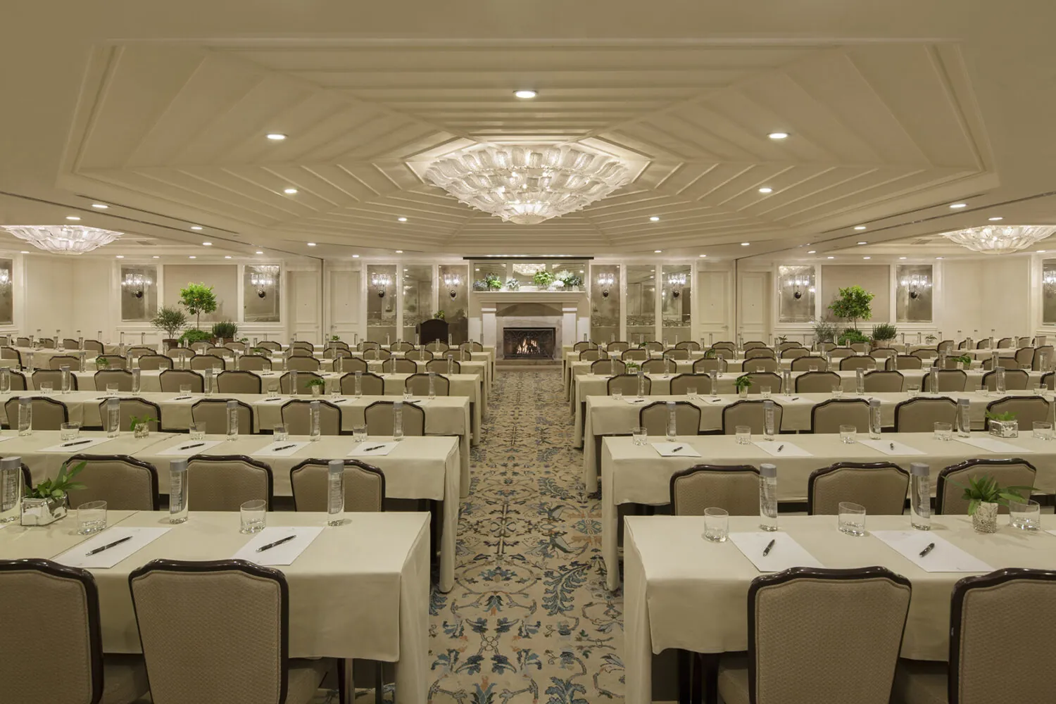 A grand conference room with elegant chandeliers, neatly arranged tables, and a warm fireplace.