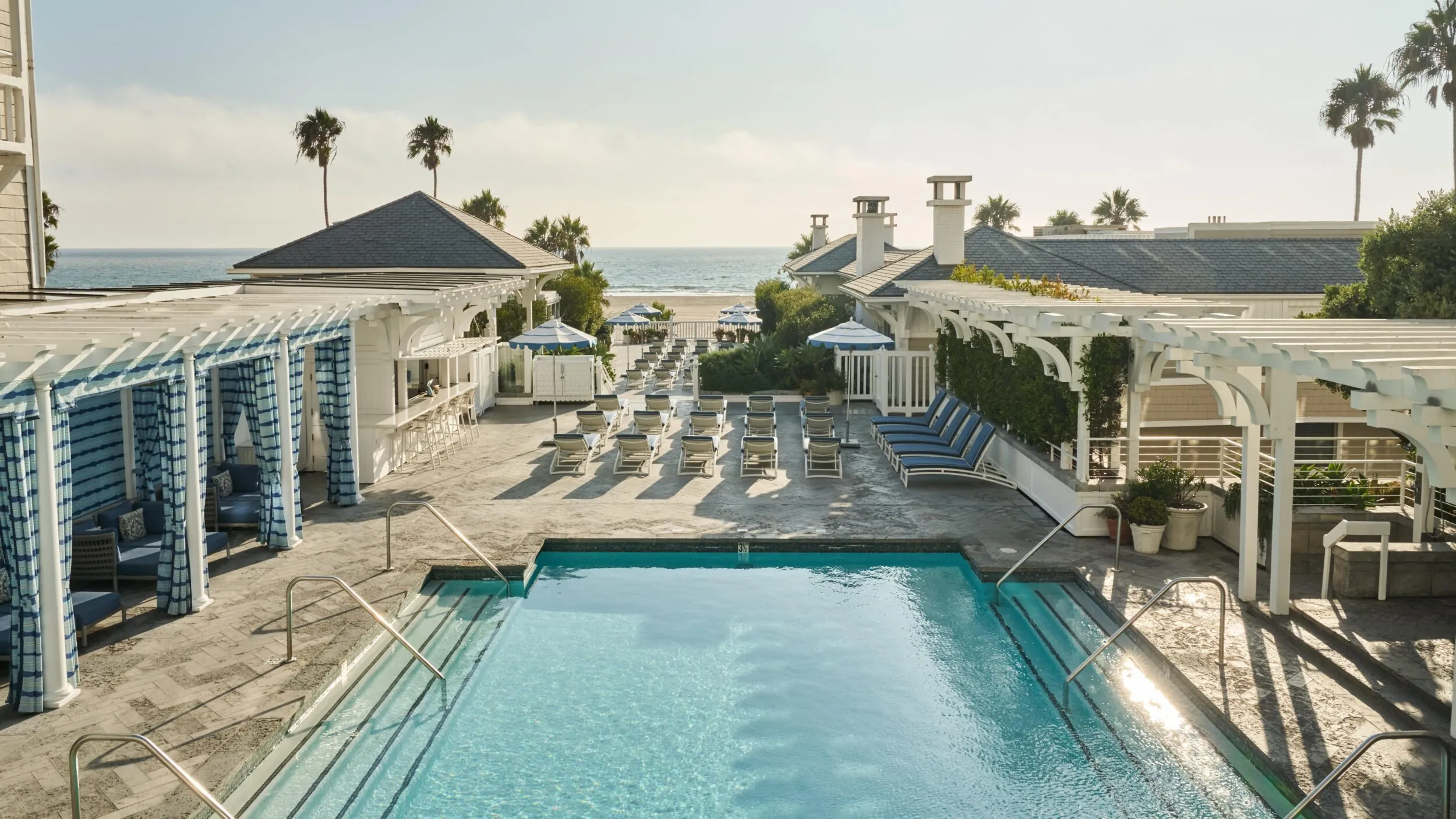Luxury beachfront pool area with lounge chairs, cabanas, and ocean views under a clear sky.