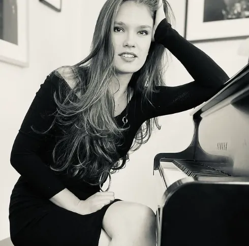 A woman in black leaning on a piano in a black-and-white photo.