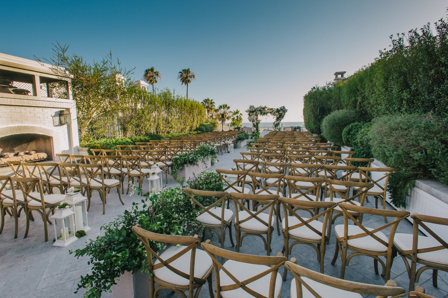 Outdoor wedding venue with rows of wooden chairs, greenery, and a clear sky.