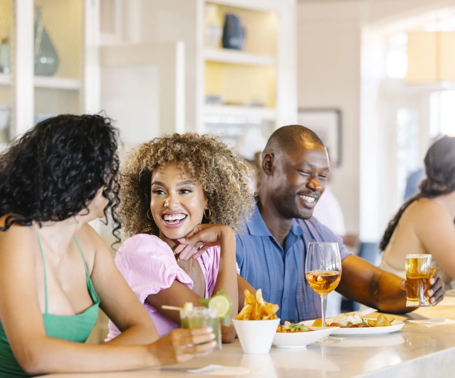 Group of friends laughing and enjoying drinks at the bar with food on the table.