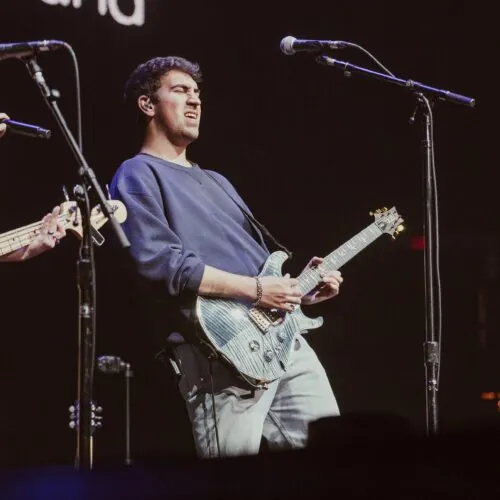 Musician playing an electric guitar on stage, wearing a blue sweater and light pants.