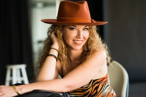 Woman with curly blonde hair, wearing a brown hat and an animal-print strapless top.