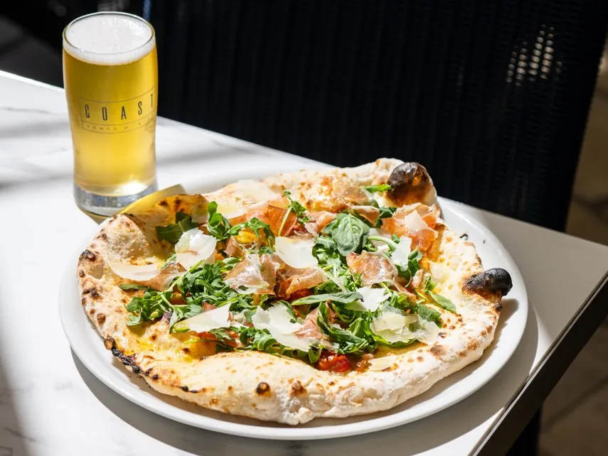 Wood-fired pizza topped with prosciutto, arugula, and a glass of craft beer on the side.