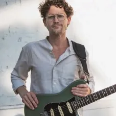 A man with curly hair and glasses holding a green electric guitar.