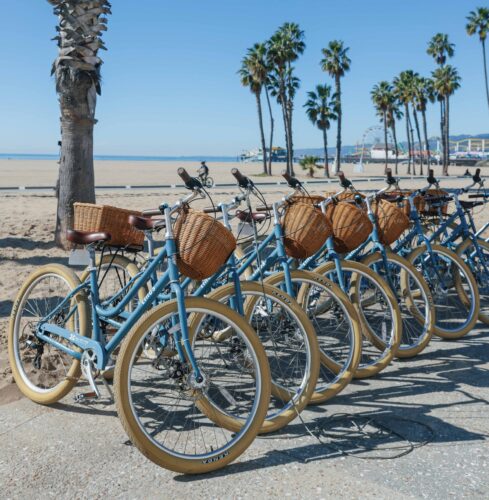 Row of light blue cruiser bicycles with wicker baskets, parked near Santa Monica beach.