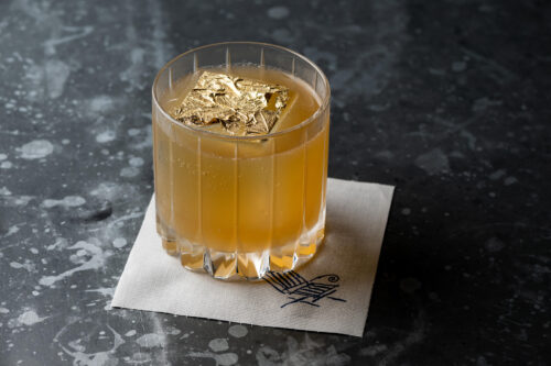 Golden cocktail in a short glass with a large gold leaf flake on top, placed on a coaster.