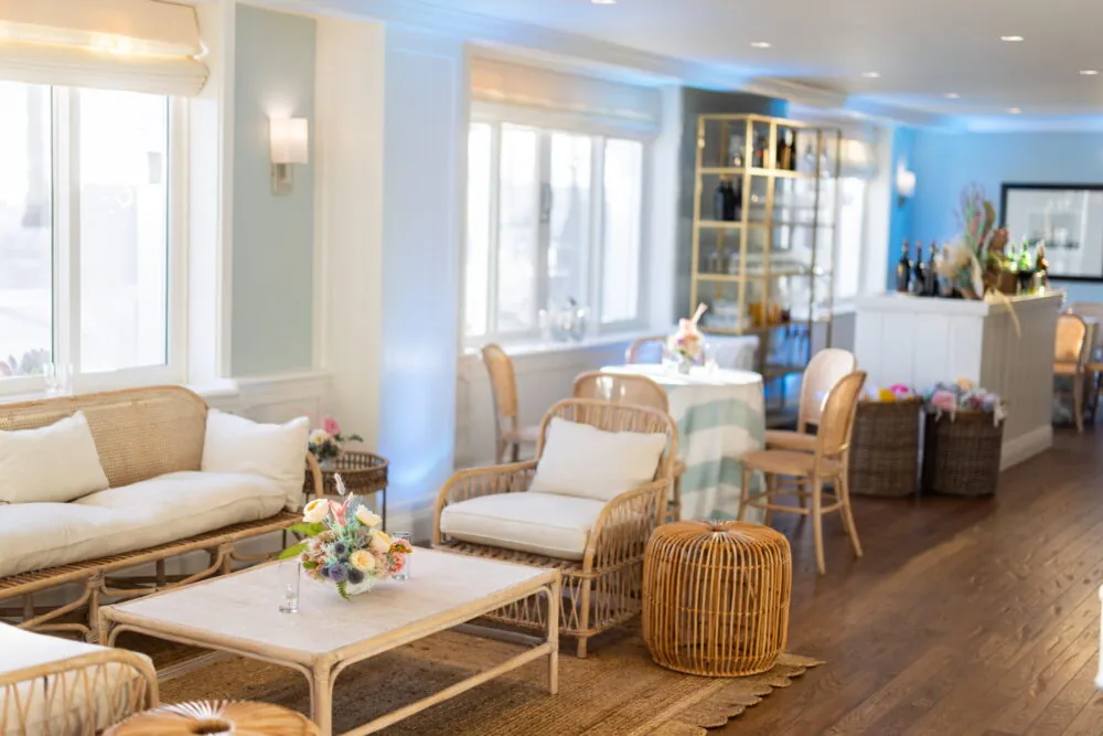 Bright, airy lounge with wicker furniture, floral centerpiece, and natural light from large windows.
