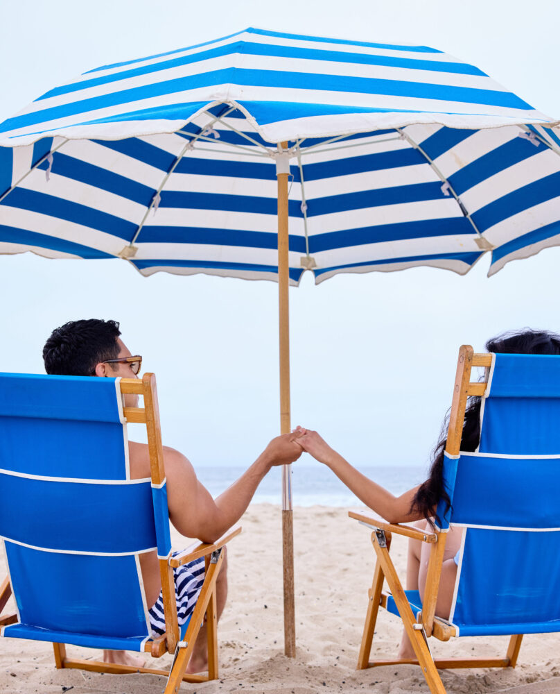 A behind view of a couple holding hands while sitting in beach chairs under an umbrella on the Santa Monica beach.