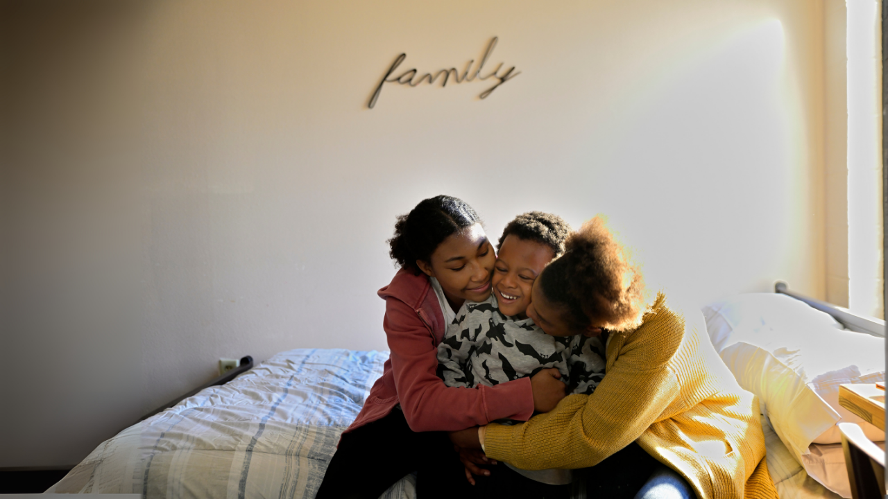 A loving family of three, two women and a young boy, embrace warmly on a bed beneath a sign that reads “family,” bathed in soft, hopeful light.