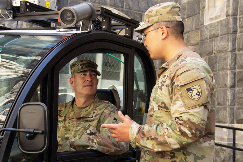 The Dean of the Academic Board, Reeves (left), sits inside the autonomous vehicle, GEM e2, as  Lim (right) explains the technological features that come with the vehicle, including the Xbox game controller for (by-wire) testing. The AVRAD team modified the vehicle installing a Linux terminal, a Light Detection and Ranging system, five Mako cameras, and configured it so that the car can be controlled through a computer. However, for safety reasons, a driver is always inside to manually control the vehicle