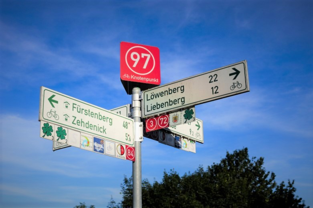 Outdoor scene featuring a tall signpost with multiple directional cycling and route signs pointing toward different destinations. The signs include distances, bicycle symbols, and route numbers, suggesting a bike trail or navigation network in a European setting.