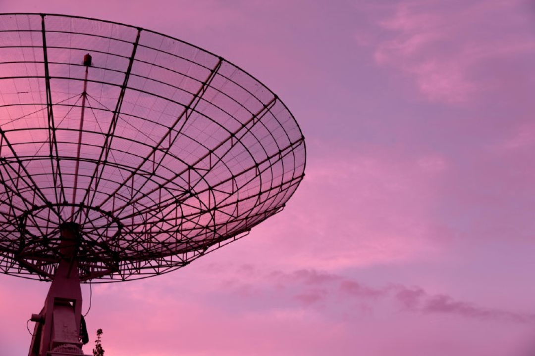A large satellite dish antenna is silhouetted against a pink and purple evening sky, representing telecommunications and signal transmission.