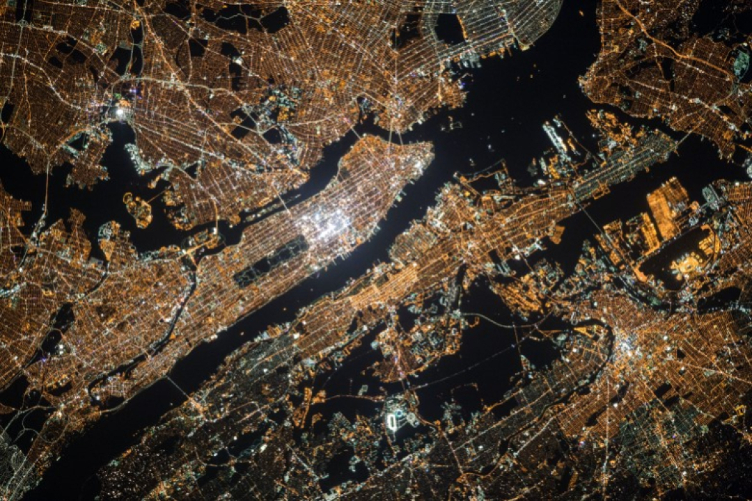 A nighttime satellite view of a densely populated city reveals glowing street grids, waterways, and illuminated urban infrastructure from above.