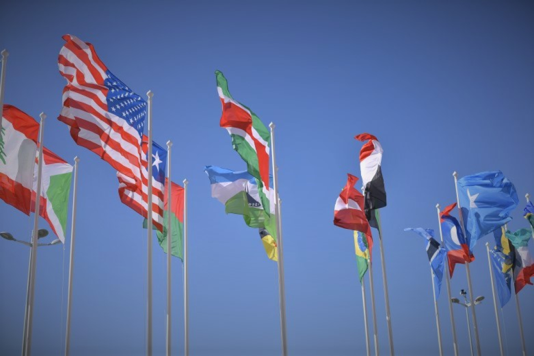 Several national flags wave on tall flagpoles against a clear blue sky, symbolizing international cooperation and global diversity.