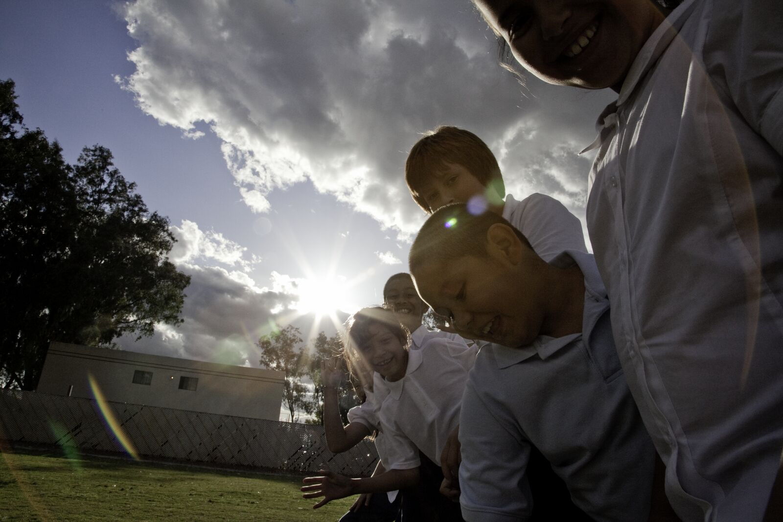 A man laughs with a group of children outdoors as the sun breaks through behind them.