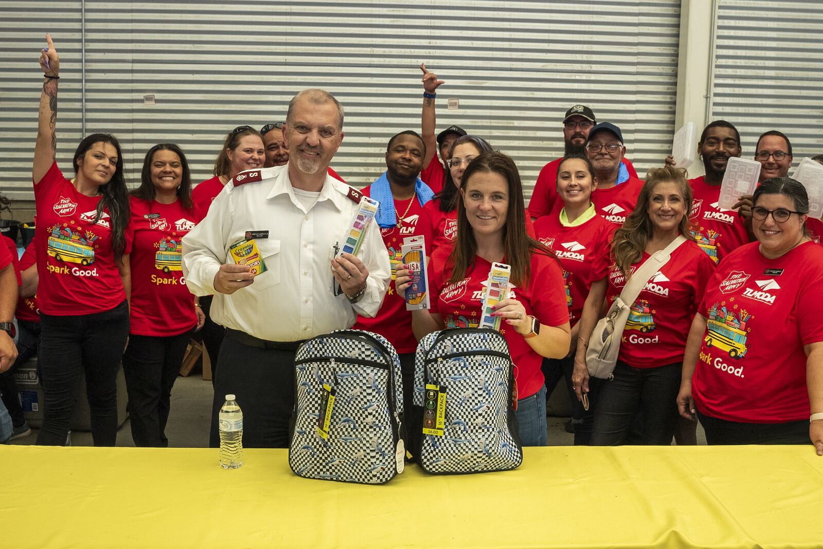A Salvation Army officer and a group of volunteers in red shirts stand behind a table of donated backpacks and school supplies.