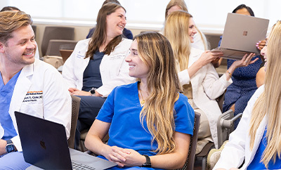 Physicians in training, some wearing scrubs and others wearing medical white coats, talk among themselves during a lecture.