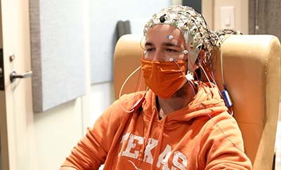 A man wearing a Texas Longhorns sweatshirt sits in a chair and wears an electroencephalogram device.