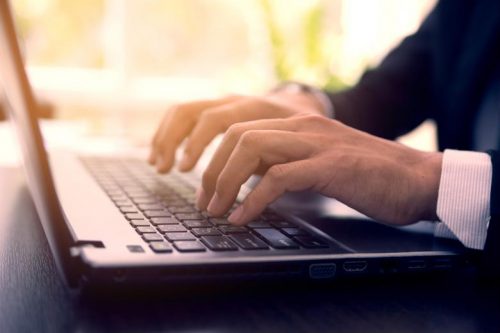 A close-up of a man’s hands typing on a laptop keyboard.