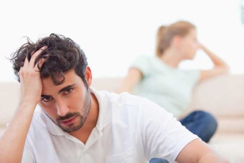 Frustrated young man with curly brown hair holding his head with his wife in the background