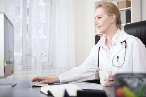 A female doctor in a white coat with a stethoscope around the neck sitting in front of her computer