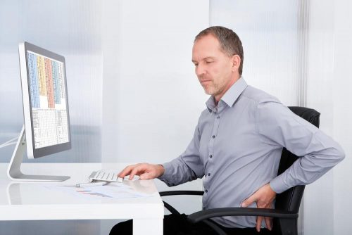 Man sitting in desk chair clutching his lower back in front of a computer, his work being interrupted by apparent back pain