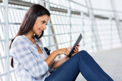 Young female with brown hair sitting on the floor against the wall while looking at the tablet