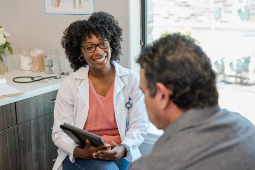 A nurse practitioner holding a tablet smiles while speaking with a patient in an exam room.