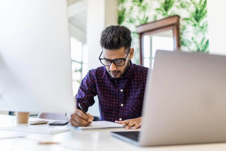 International Tax Manager writing on a notepad behind desktop and laptop computers