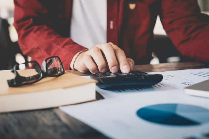 Close up view of a tax accountant typing into a calculator with books, paper graphs and glasses on the desk.