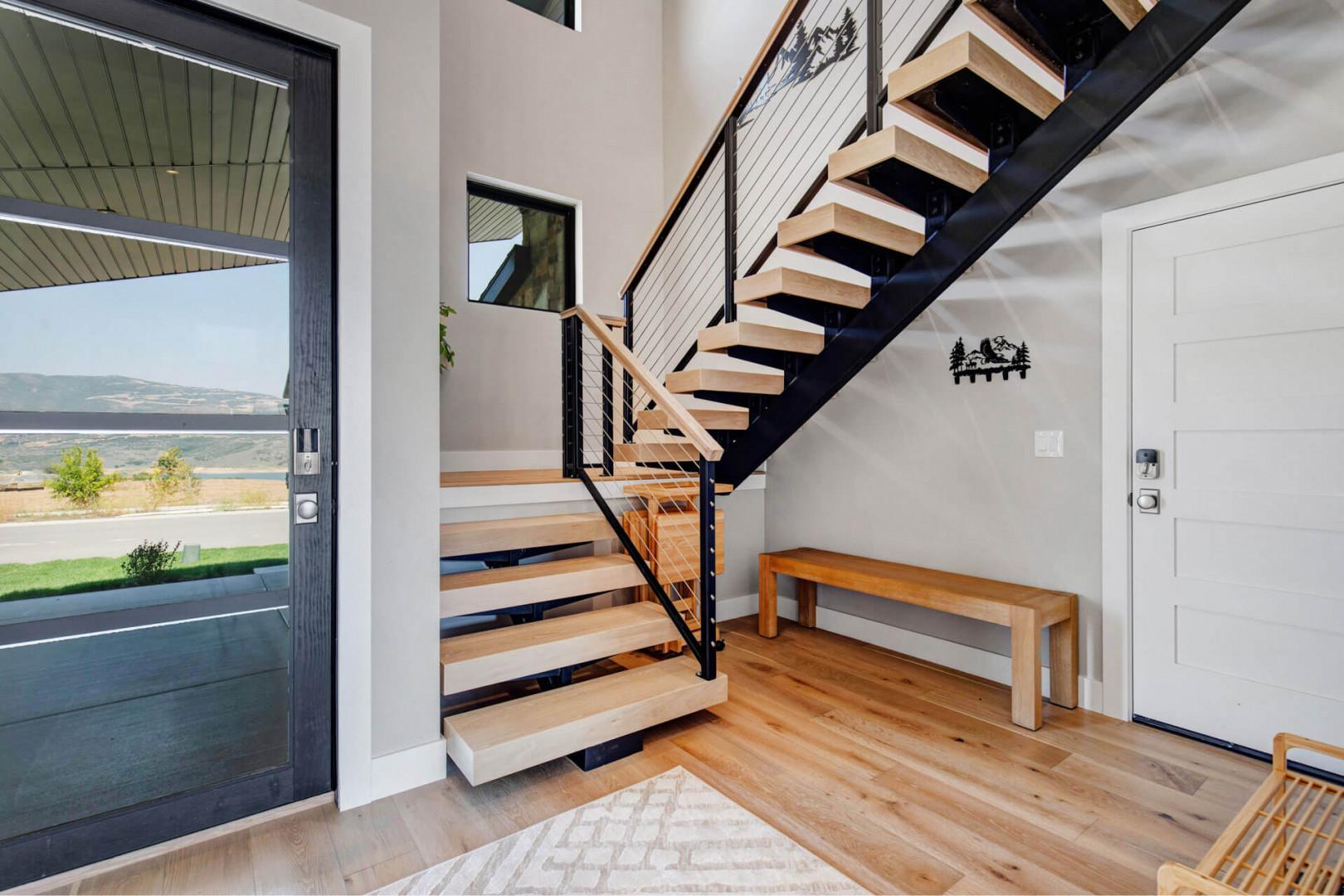 Modern entryway with striking open staircase and natural light streaming through the glass door, inviting you into your mountain retreat with warm wood floors beneath your feet.