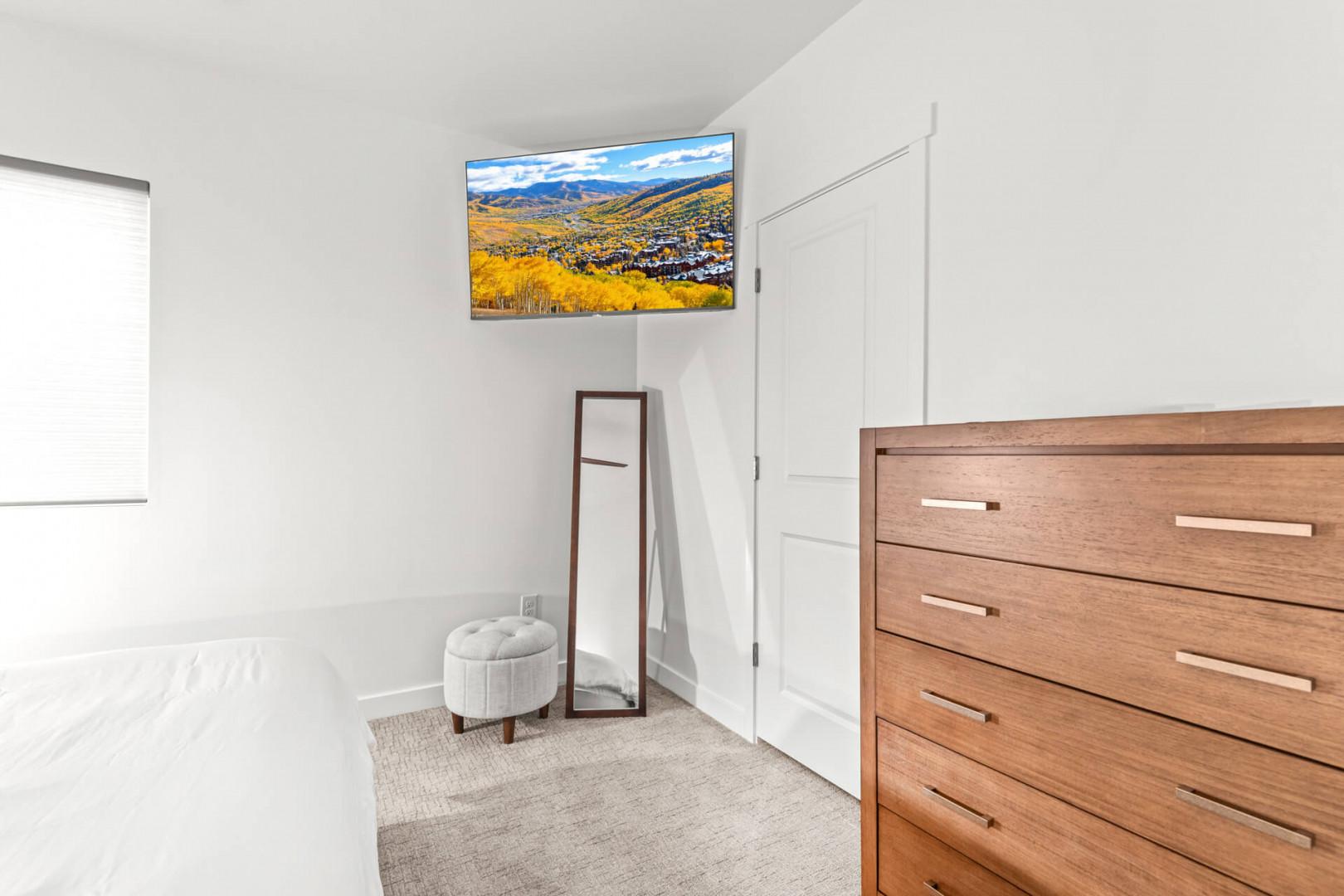 Relax in your serene bedroom with a wall-mounted TV showcasing stunning mountain views. Soft natural light and modern furnishings create a peaceful retreat for restful evenings.