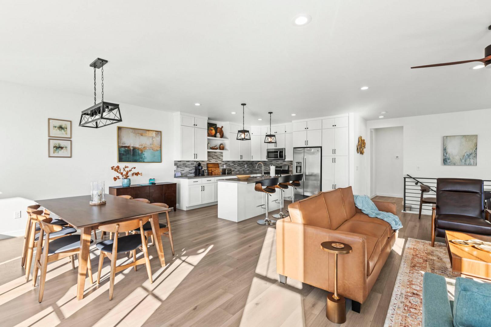 Unwind in your open-concept living space where modern kitchen, dining, and lounge areas flow together. Sunlight pours across warm wood floors, highlighting sleek white cabinetry and inviting leather seating—your home away from home.