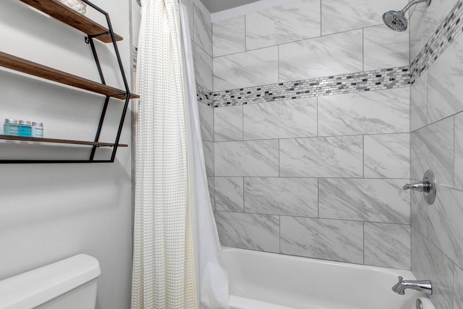 Refresh in your stylishly tiled bathroom featuring a marble-look shower-tub combo with modern fixtures and convenient shelving for all your essentials.