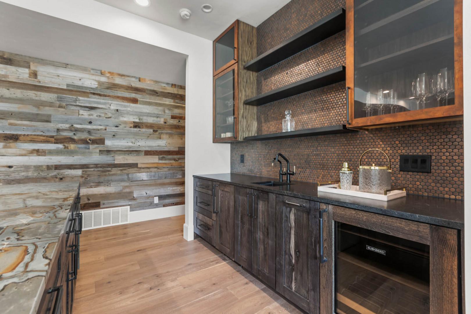 Your stylish wet bar features rustic wood cabinetry with glass doors, elegant penny tile backsplash, and dark countertops. Stocked with glassware and ready for entertaining with modern appliances.