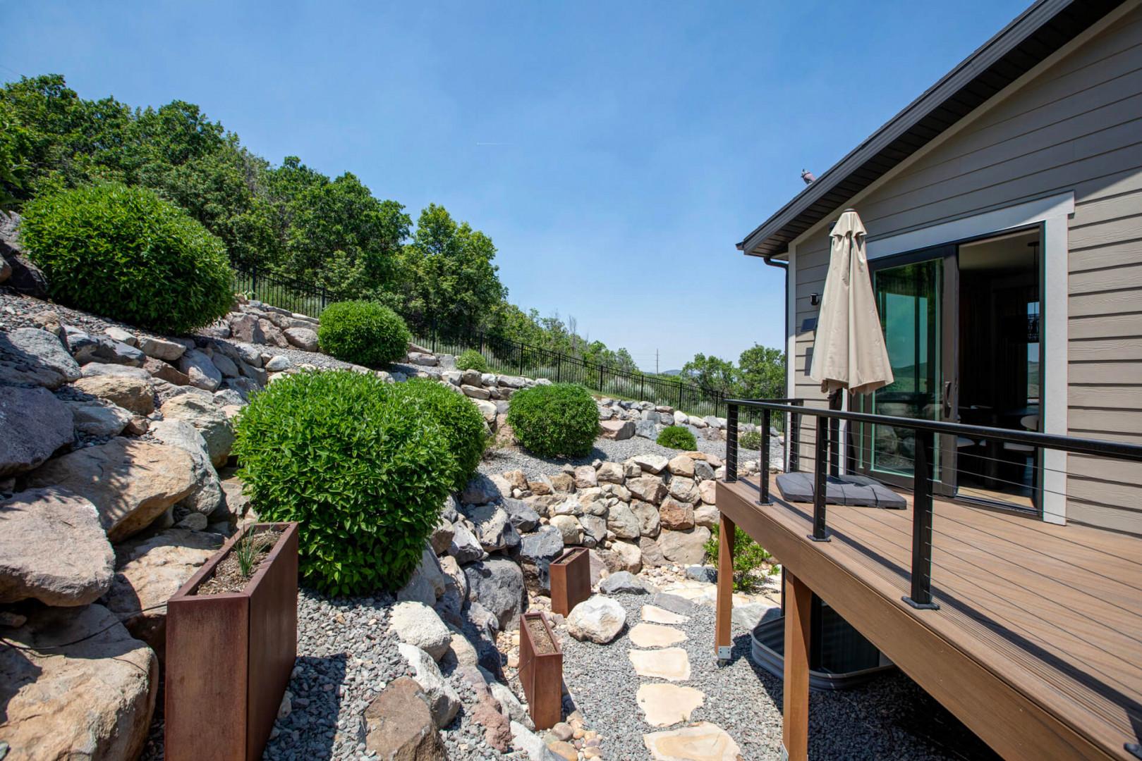 Modern rental situated on a natural hillside with landscaped rock gardens and lush greenery surrounding the property, featuring contemporary exterior design and elevated deck space.