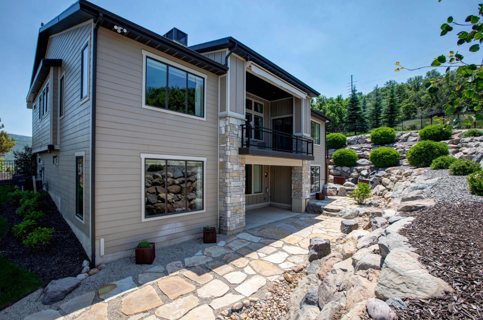 Modern two-story vacation home nestled against a landscaped hillside, with natural stone pathways leading to covered balcony and private entrance.