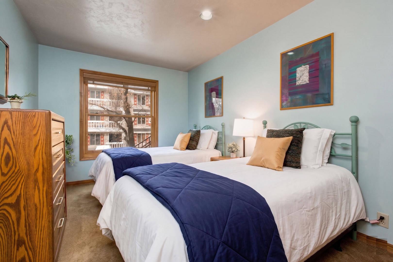 Step into your peaceful bedroom retreat with two plush beds dressed in crisp linens and navy accents, where soft light and calming blue walls invite you to unwind after your day of adventure.