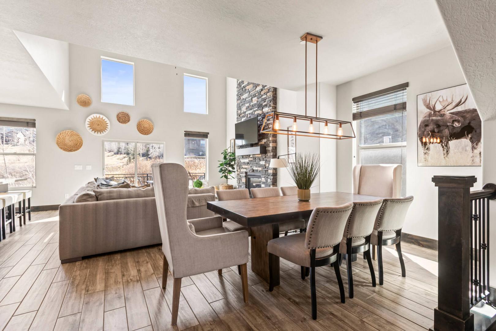 Gather around a handsome wood table under natural light streaming through expansive windows, with mountain views and a cozy stone fireplace setting the scene for memorable meals together.
