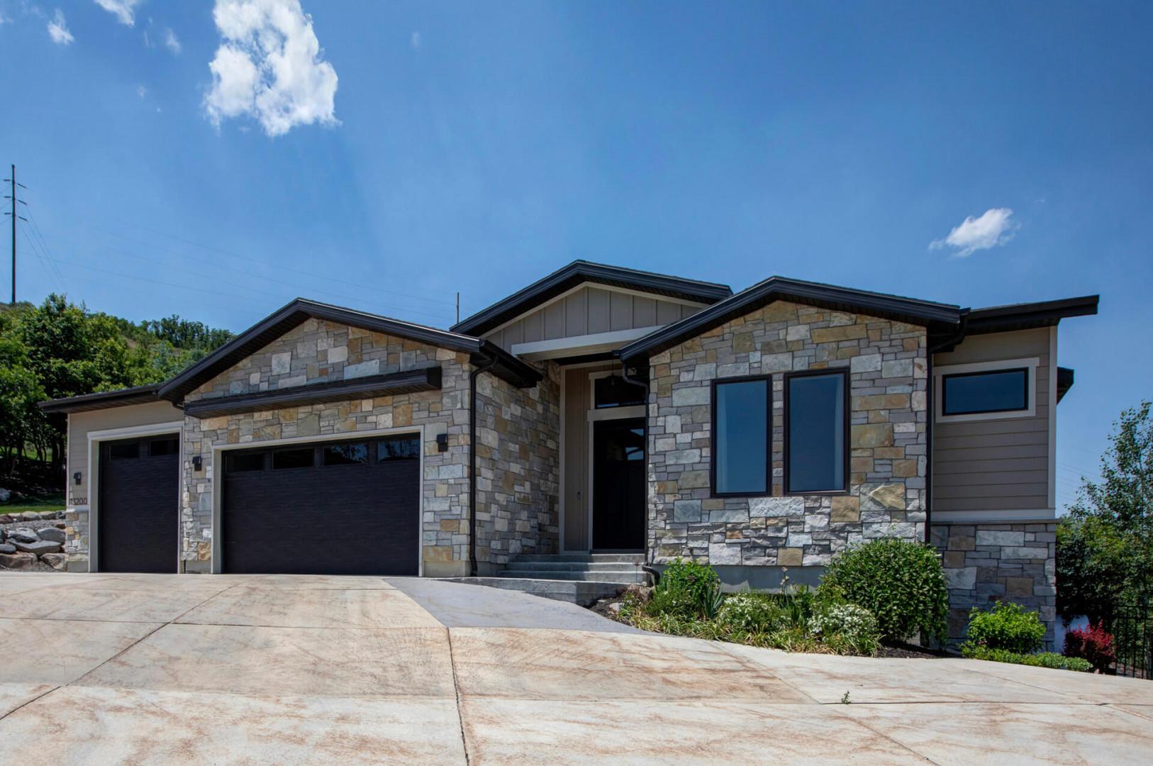 Modern stone-and-siding vacation home nestled among trees with spacious driveway parking and sunny skies, welcoming guests to a peaceful hillside retreat.