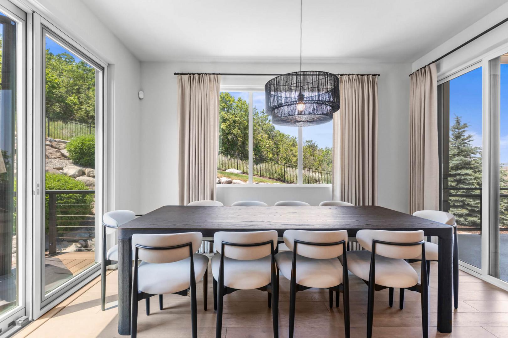 Gather around this elegant dining table where natural light pours in through expansive windows, framing views of lush greenery and creating the perfect setting for memorable meals together.