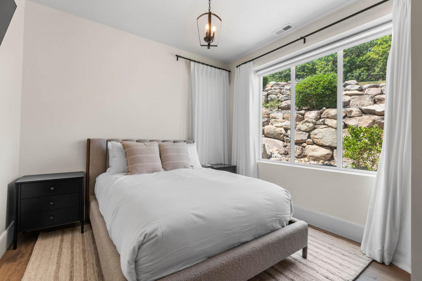 Sink into crisp linens in your serene bedroom, where natural light frames a peaceful stone-wall view and soft curtains enhance your restful escape.