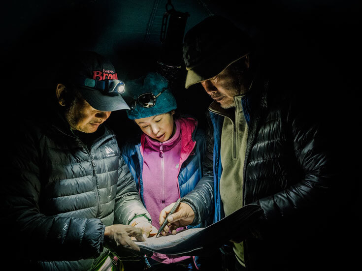 A woman stands between two men, wearing jackets and hats and standing in darkness as they consult a map.