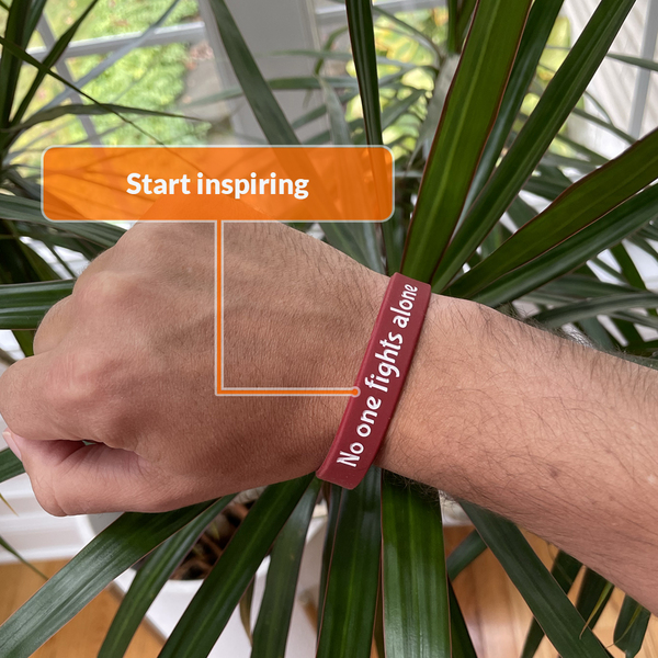 dark red wristband that says no one fights alone being worn on a wrist with the background of a plant